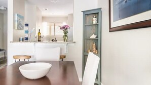 A dining area with a wooden table, white chairs, and a white bowl. There is a kitchen in the background with a window, a vase with flowers, and a cabinet with decorative items.