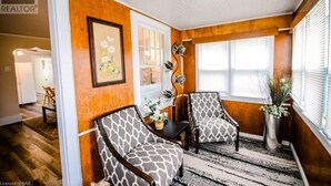 Una habitación con dos sillas estampadas, una obra de arte enmarcada, un jarrón con flores y una ventana con persianas.