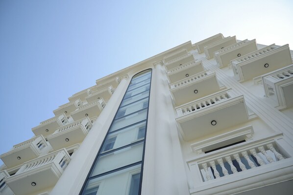 Un edificio de varios pisos con un elevador central de vidrio, balcones blancos y un cielo despejado.