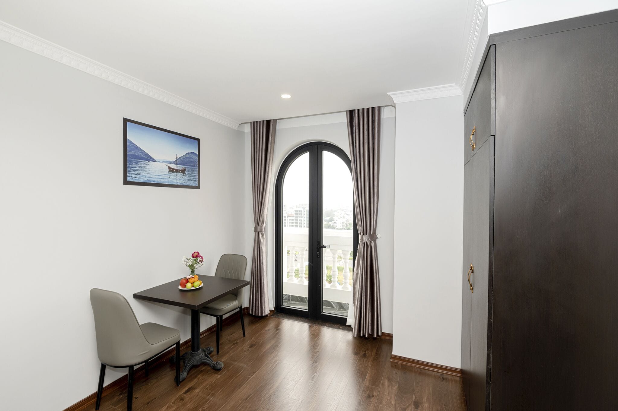 A room with a dining table, two chairs, a vase with flowers, a painting, and a view through a large window with curtains.