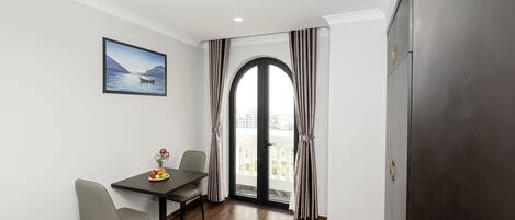A room with a dining table, two chairs, a vase with flowers, a painting, and a view through a large window with curtains.