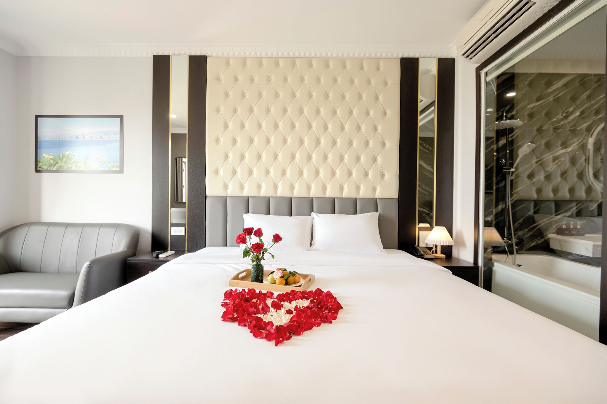 A hotel room with a large bed, a grey sofa, a bedside table with a vase of flowers, and a bathroom with a marble tub.