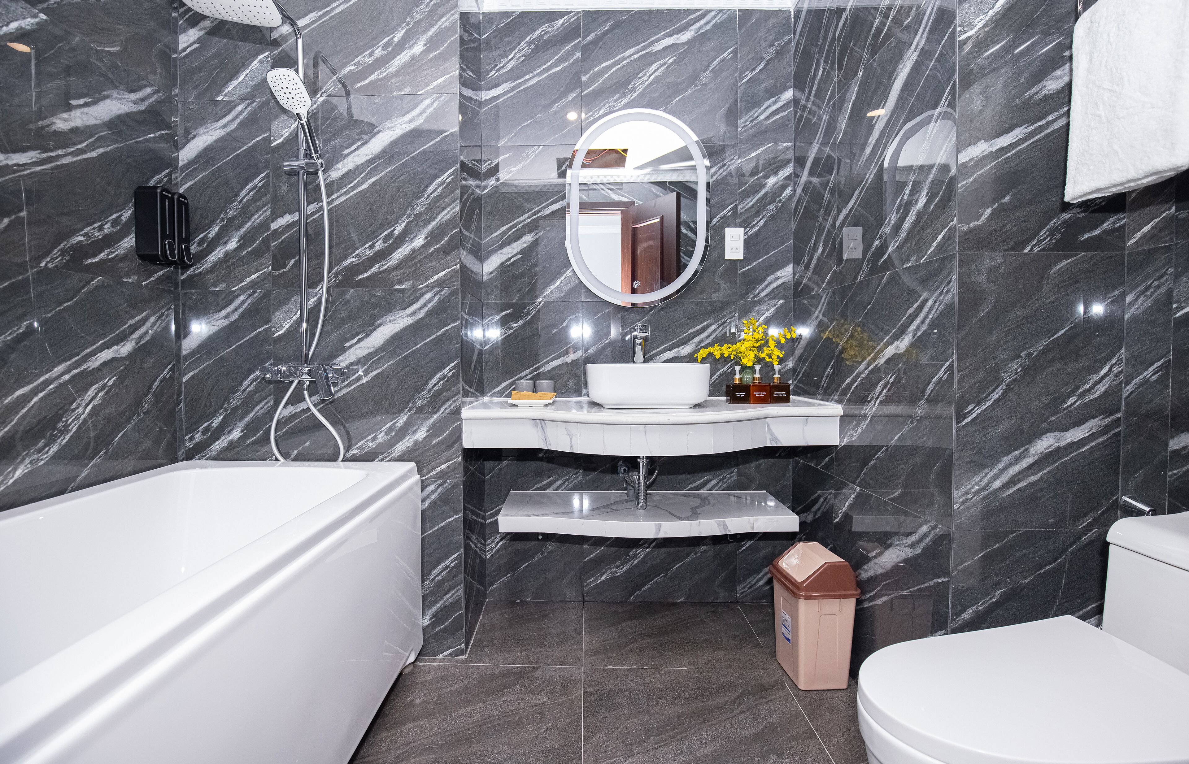 A modern bathroom with a marble wall, a white bathtub, a sink with a mirror, and a toilet.