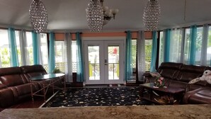 Ein Wohnzimmer mit braunem Ledersofa, einem Couchtisch und einem Esstisch mit Stühlen. Zwei große Fenster mit Vorhängen, eine zur Außenwelt führende Tür und eine Chandelier.