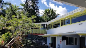 Ein zweistöckiges Gebäude mit Balkon, umgeben von tropischer Vegetation und blauem Himmel.