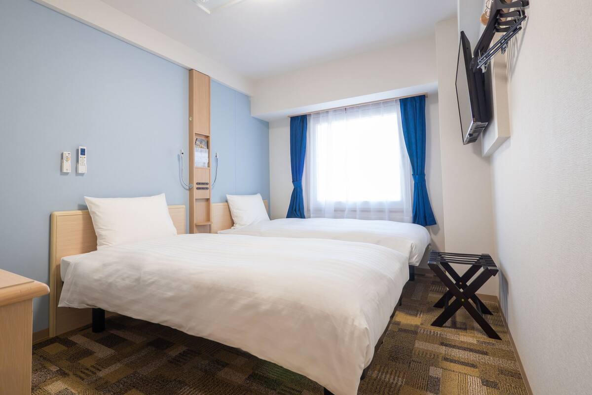 A hotel room with two beds, a desk, a chair, a TV, and a window with curtains.