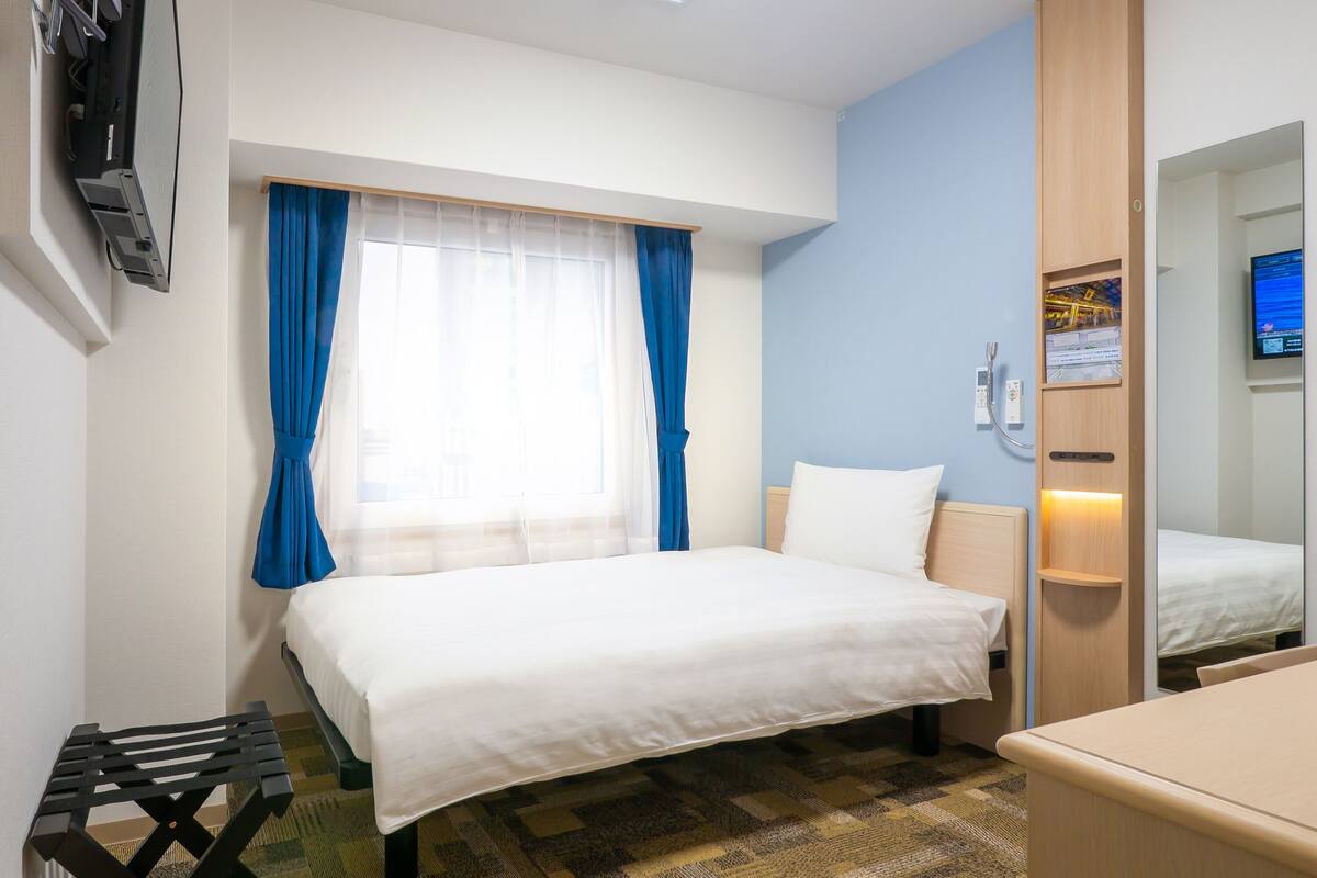 A hotel room with a bed, a desk, a chair, a window with curtains, a mirror, and a wall-mounted TV.