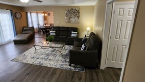 Una sala de estar con sofá, sillón y mesa de centro. En el fondo, se observa un comedor con una mesa y sillas.