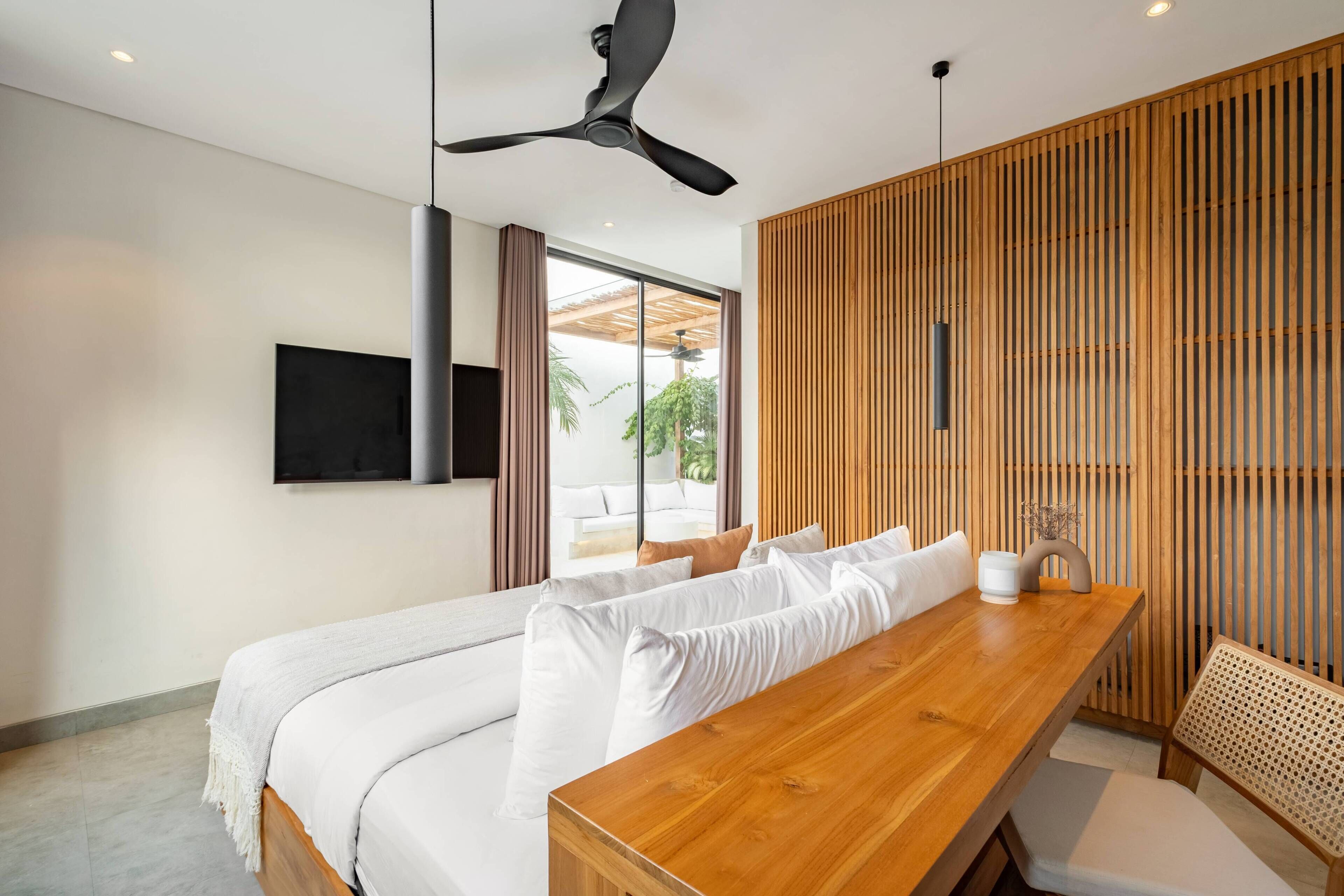 A modern hotel room with a wooden dining table, a sofa with white cushions, a ceiling fan, and a TV mounted on the wall.