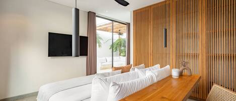 A modern hotel room with a wooden dining table, a sofa with white cushions, a ceiling fan, and a TV mounted on the wall.