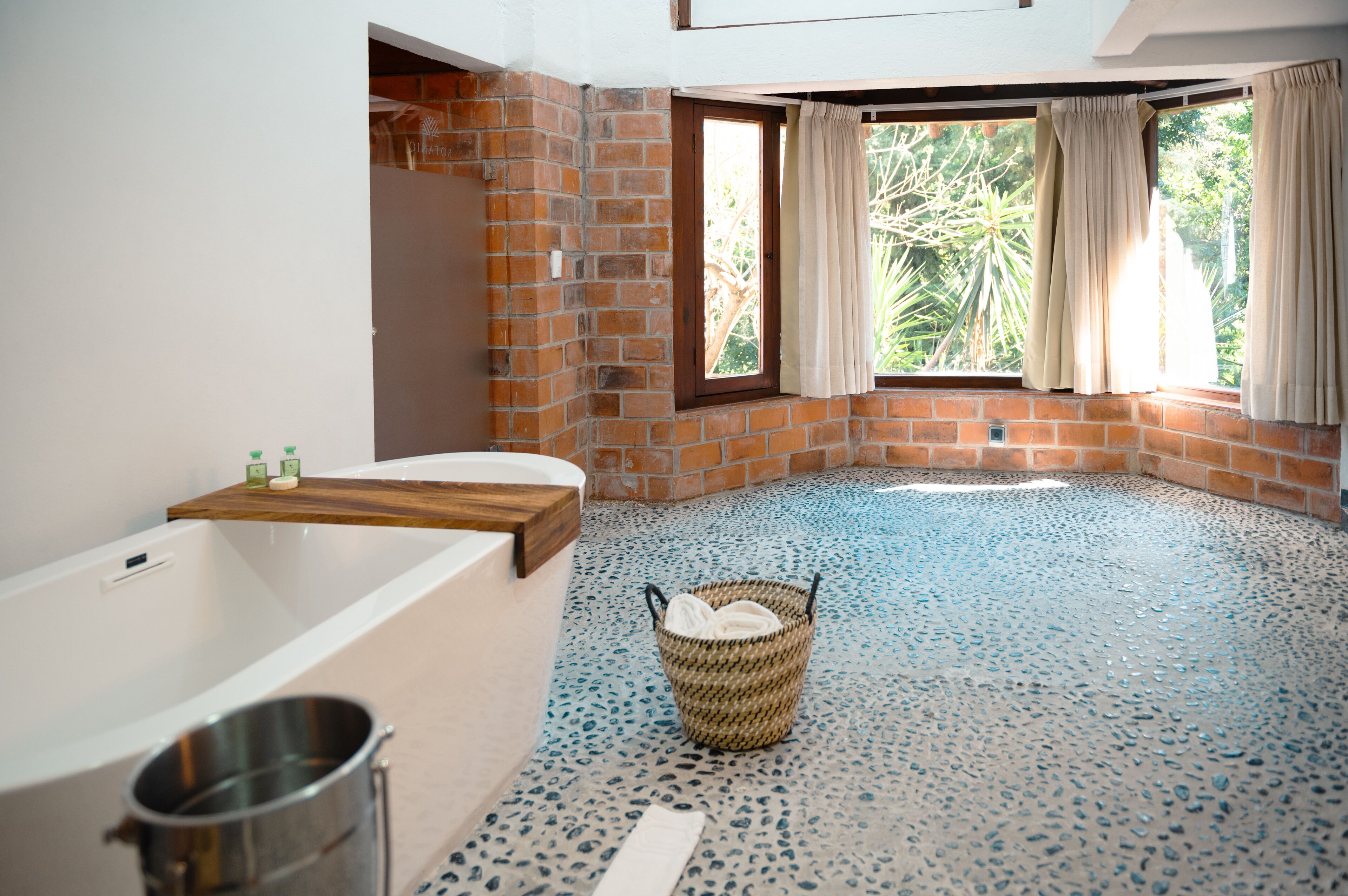 A bathroom with a bathtub, a window with curtains, a tiled floor, and a wicker basket.