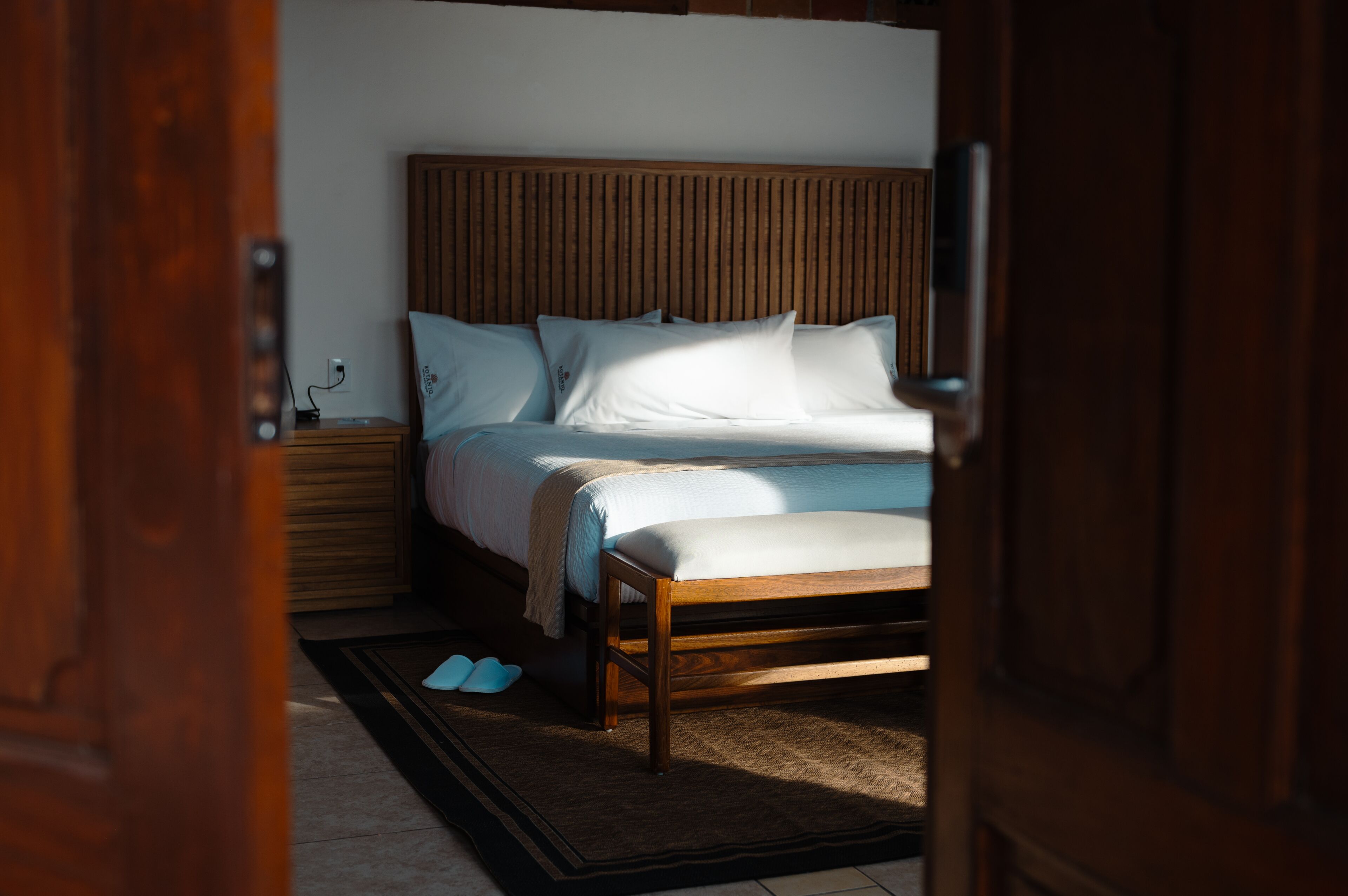 Un dormitorio con cabecera de madera, cama con ropa de cama blanca, mesita de noche con lámpara y un par de zapatillas en el suelo.