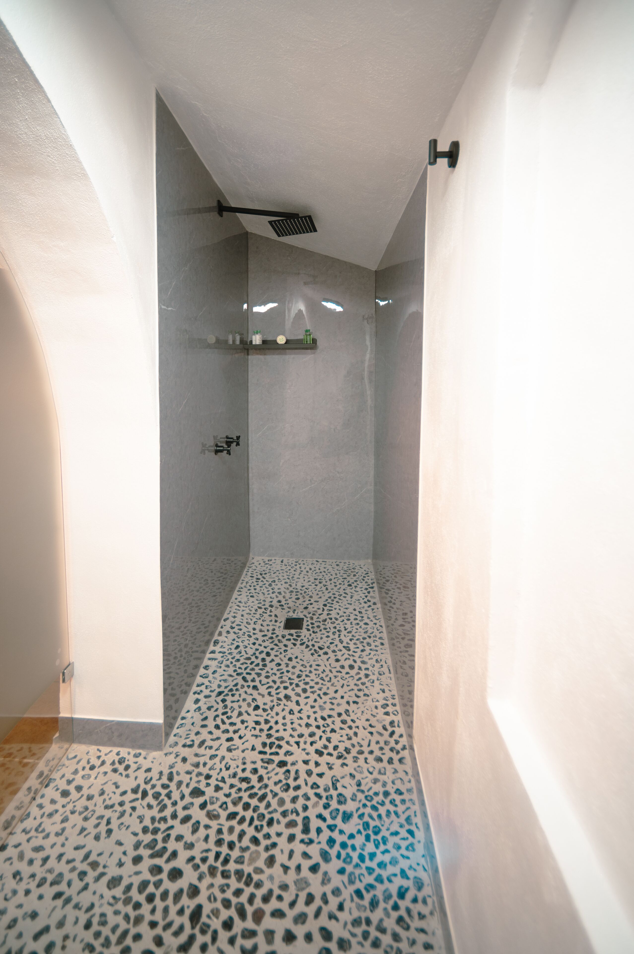 A modern shower area with a speckled floor, a ceiling-mounted showerhead, and a shelf with toiletries.