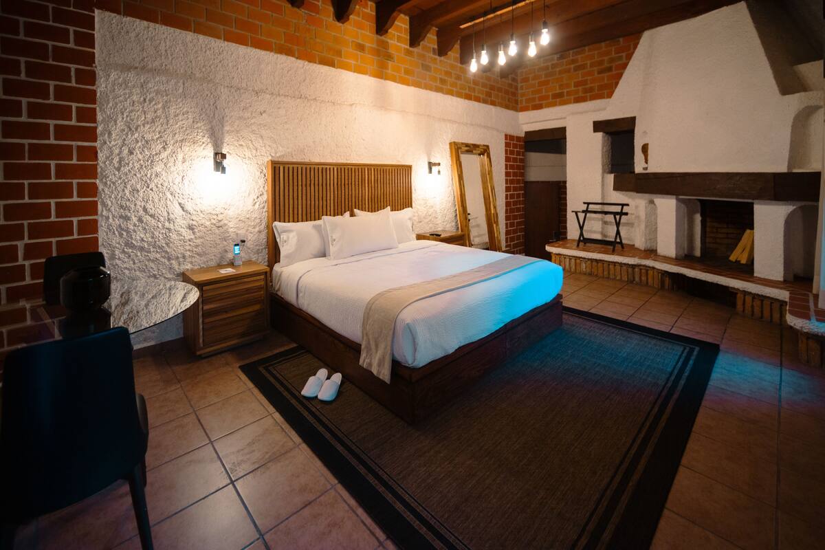 A bedroom with a large bed, a nightstand, a chair, and a fireplace.