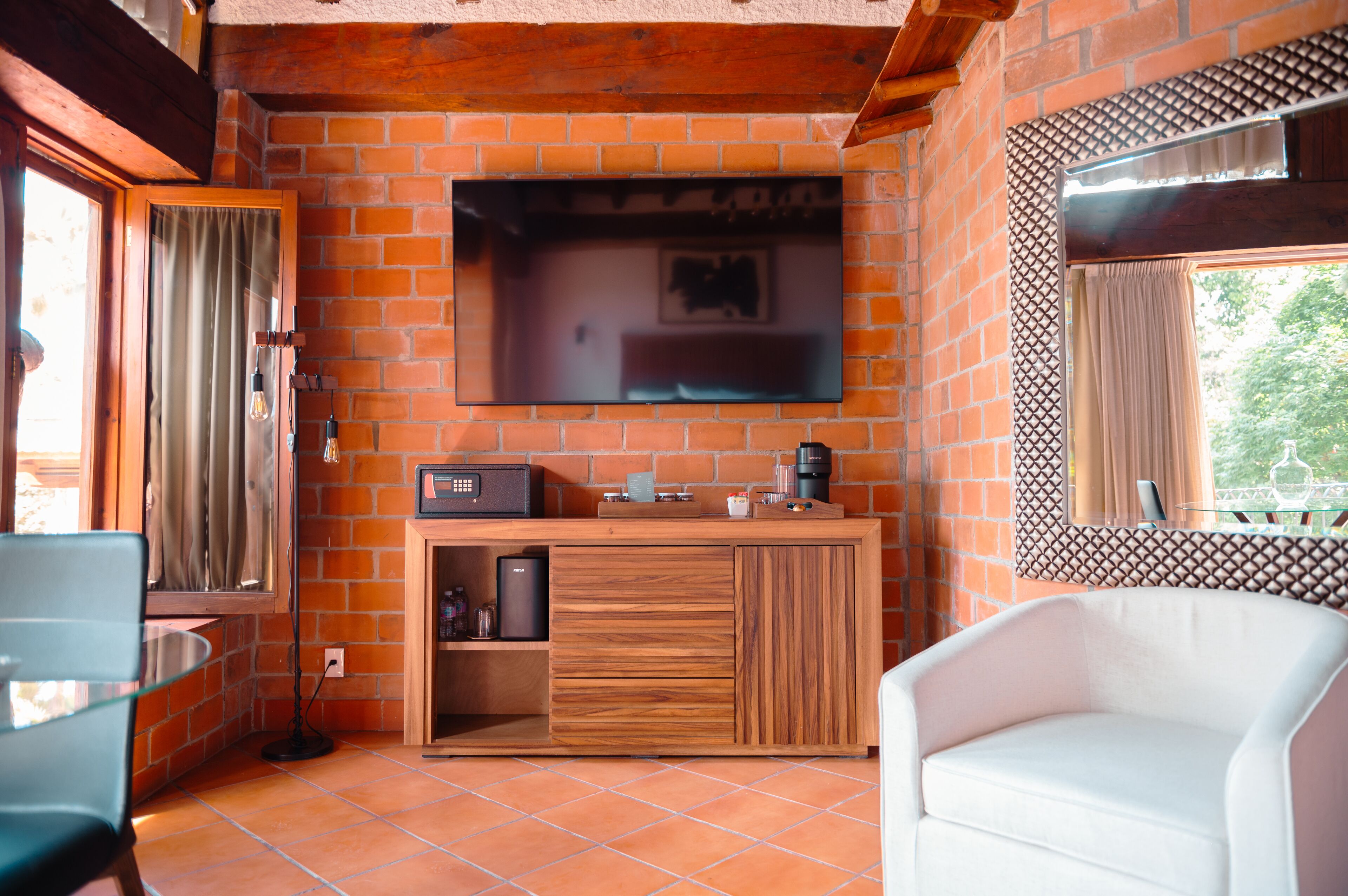 A cozy room with a brick wall, a flat-screen TV mounted above a wooden counter, a white sofa, and a window with a view of greenery.