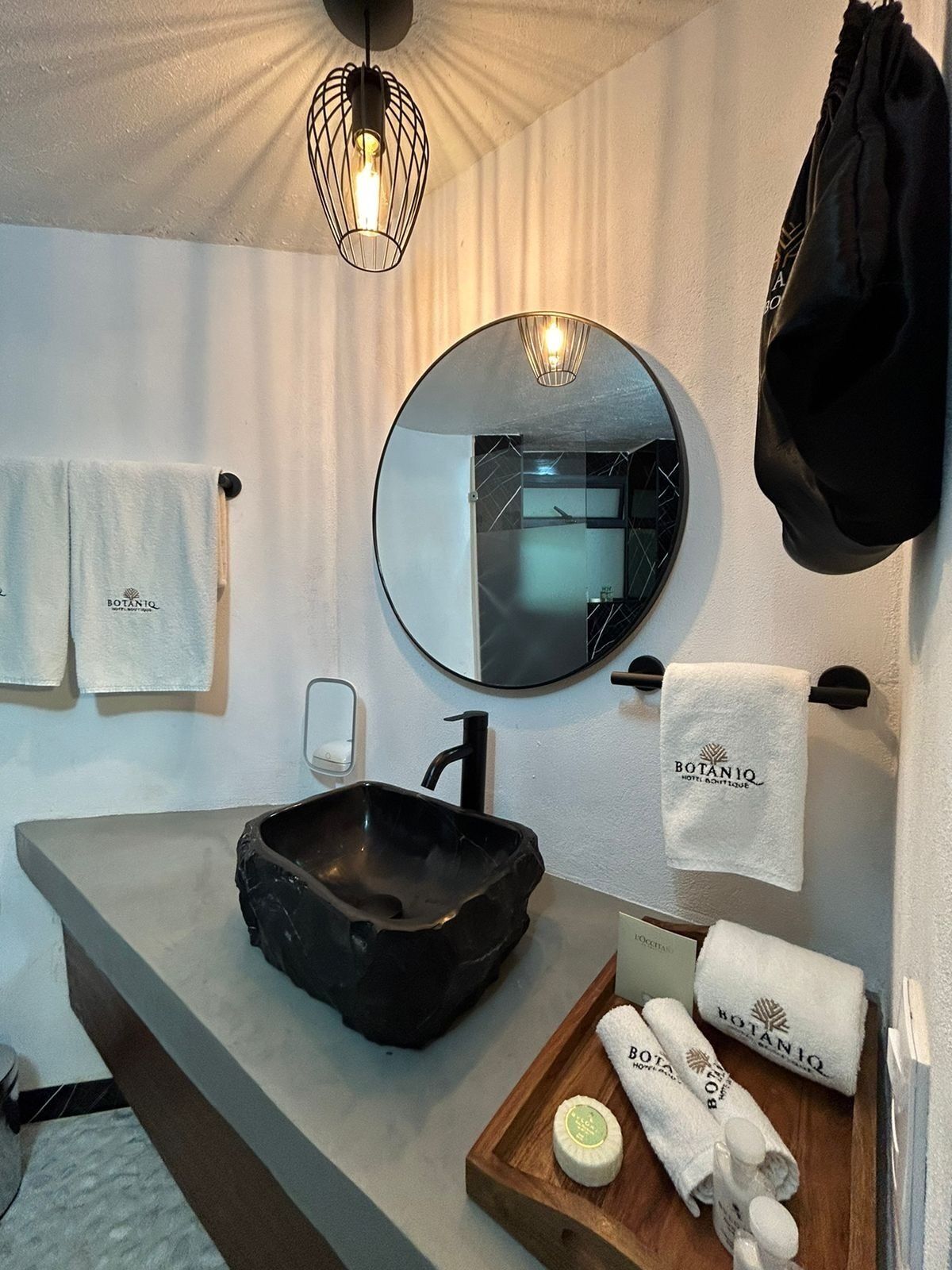 A bathroom with a black sink, a round mirror, a towel rack, and a wooden tray with toiletries.
