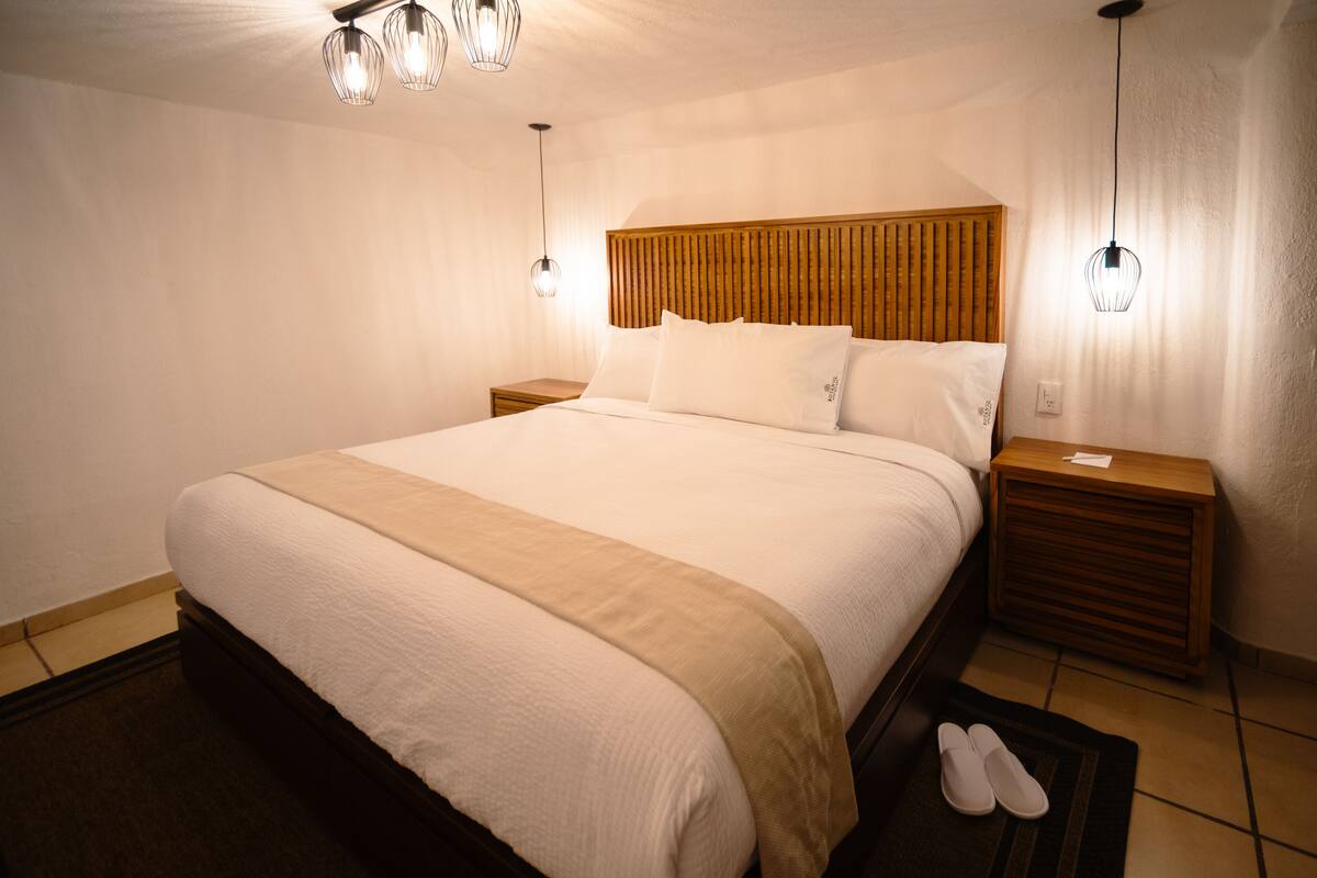 A neatly made bed with a wooden headboard, two bedside tables, and a pair of slippers on a rug.