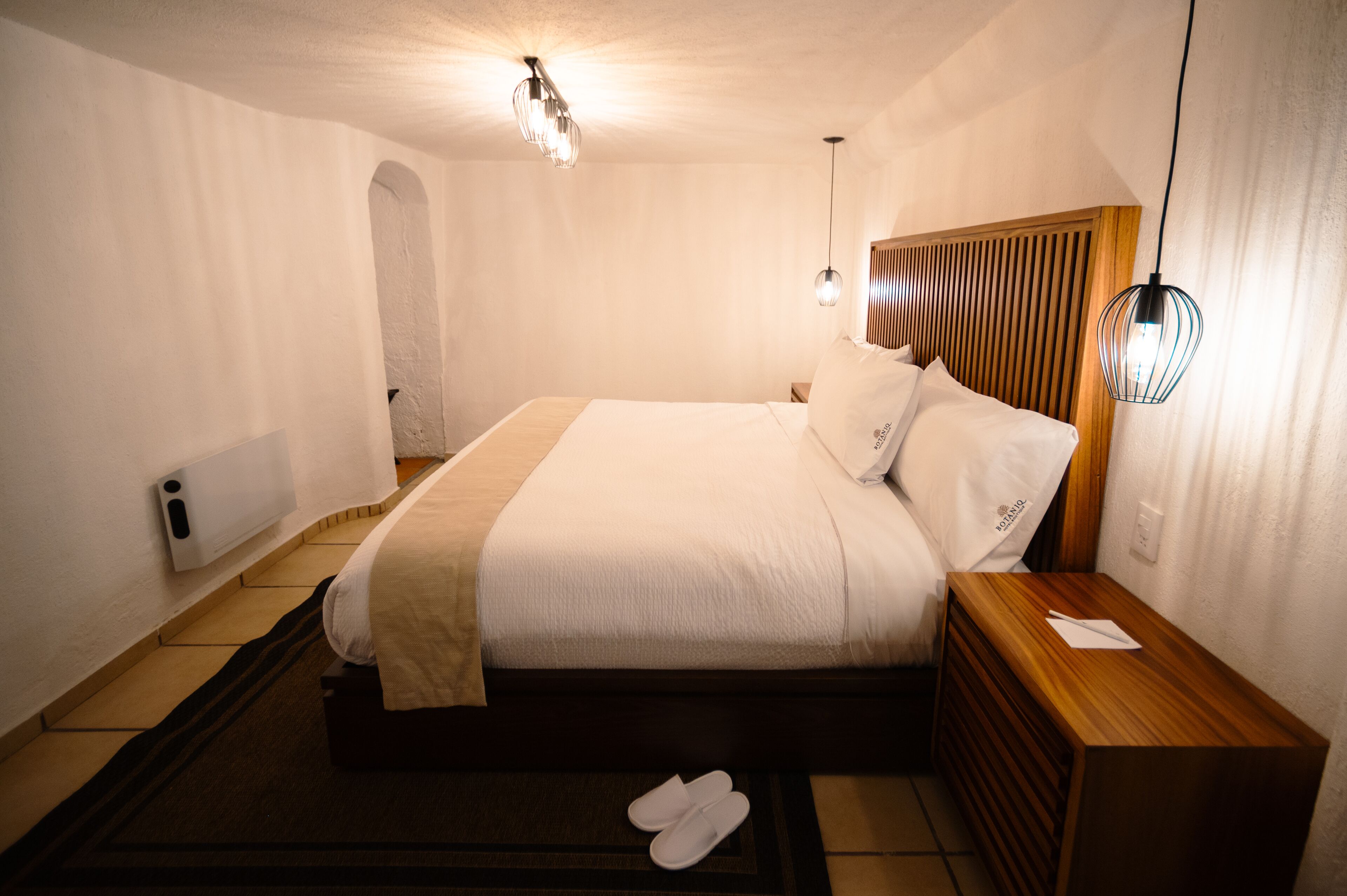 A hotel room with a bed, nightstand, and a pair of slippers on the floor.
