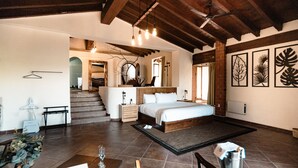 Un dormitorio espacioso con una cama grande, cabecera de madera y vistas a una escalera. Se observa una mesita de noche con una lámpara y una pequeña planta. El cuarto tiene piso de baldosas y un ventilador de techo.