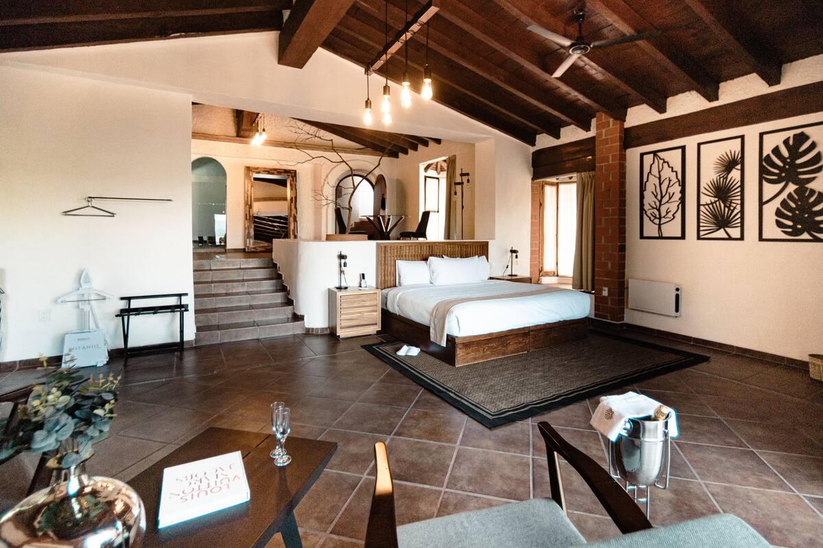 A spacious bedroom with a large bed, a wooden headboard, and a view of a staircase. There is a bedside table with a lamp and a small plant. The room has a tiled floor and a ceiling fan.