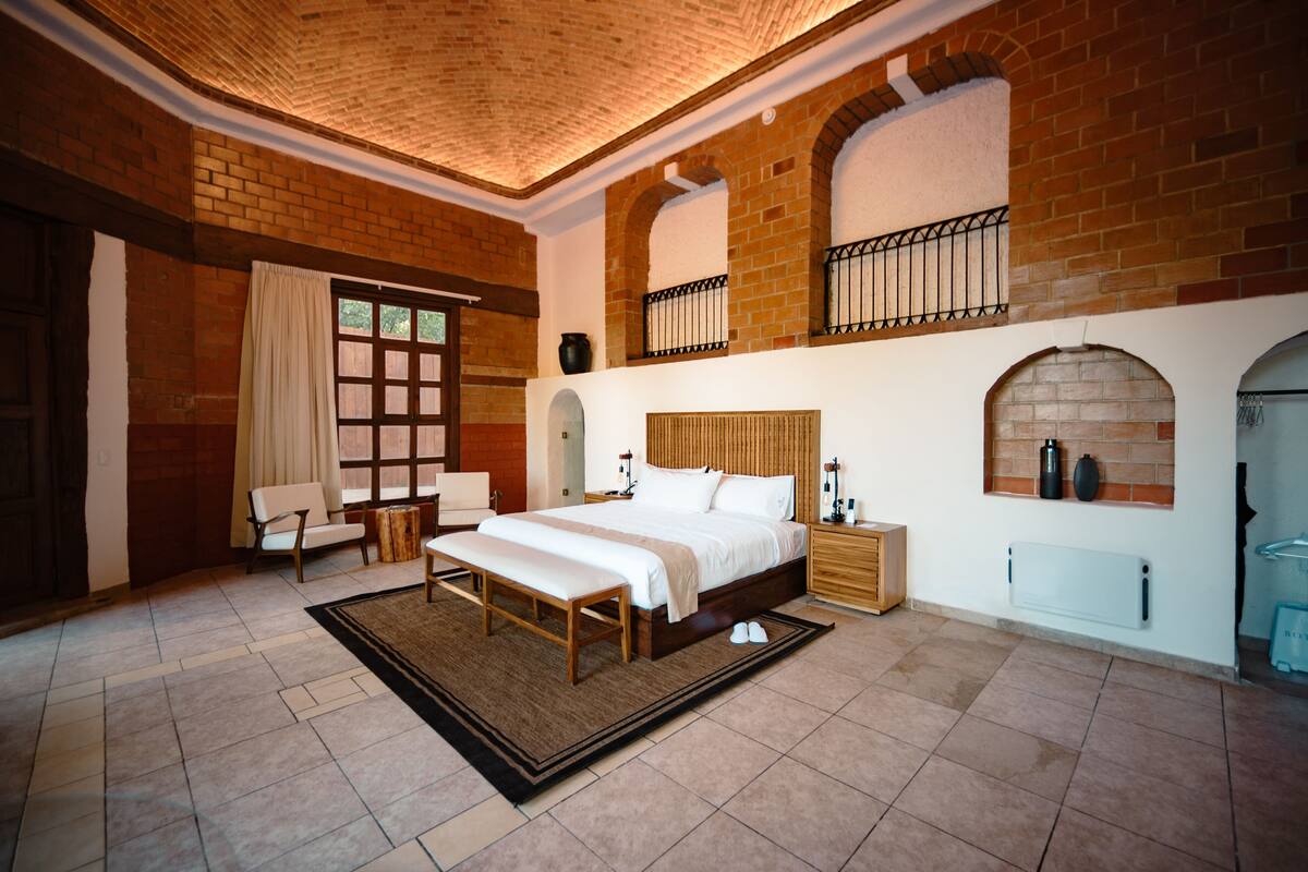 A bedroom with a large bed, a wooden bench, a nightstand, and a brick wall with arched windows.