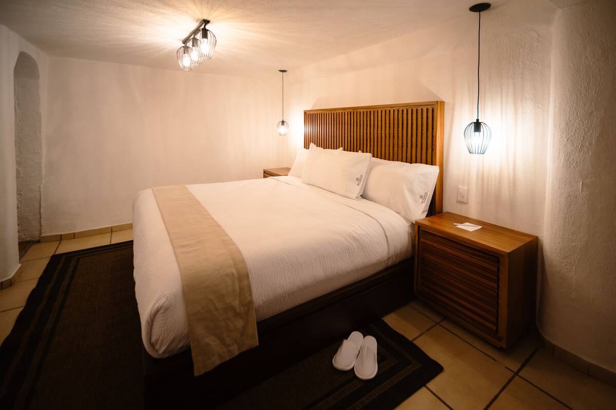 A neatly made bed with a wooden headboard, a bedside table with a lamp, and a pair of slippers on the floor.