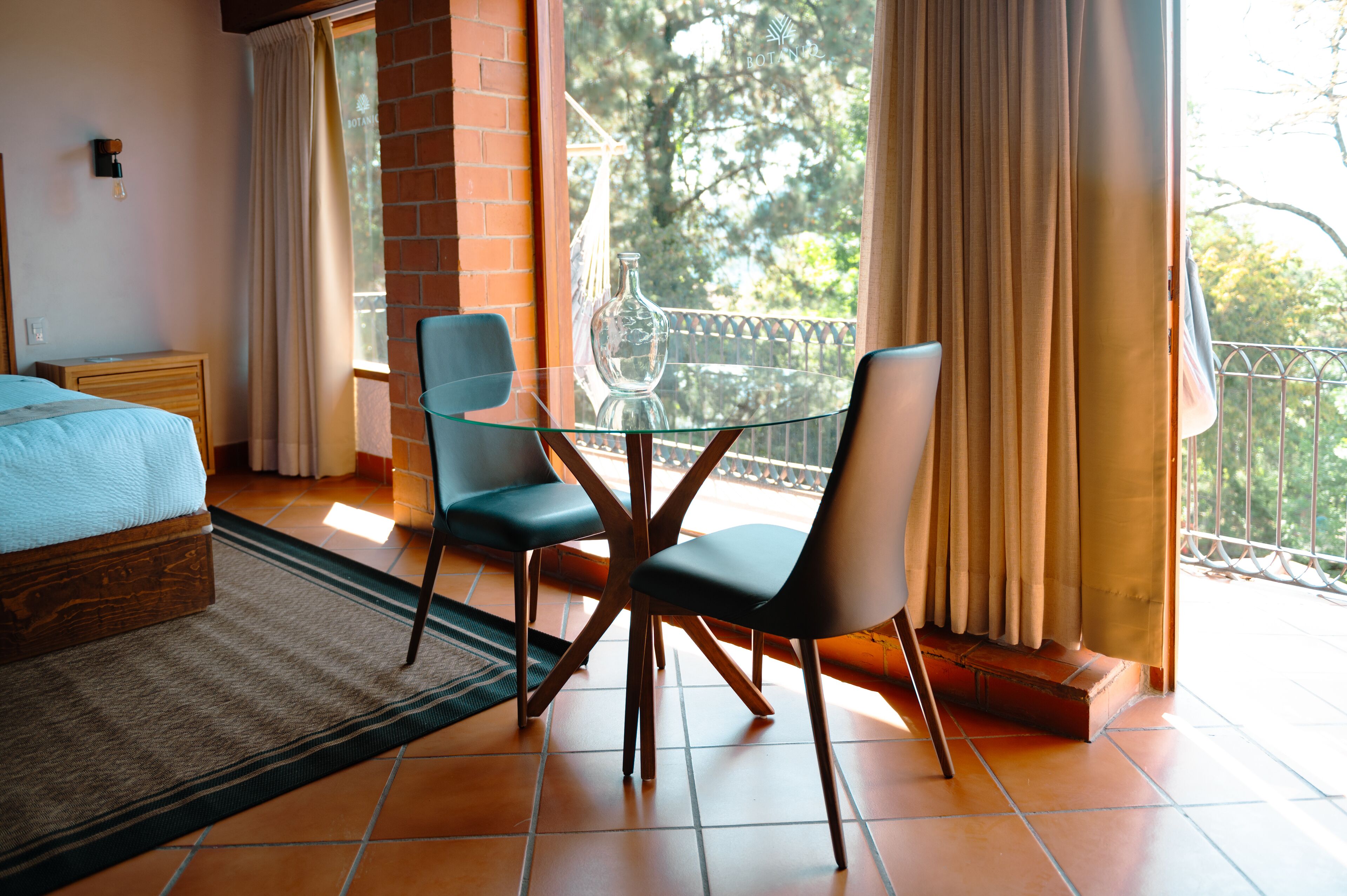 Un dormitorio con cama, una mesa de vidrio y sillas, una ventana con cortinas y vistas a árboles.