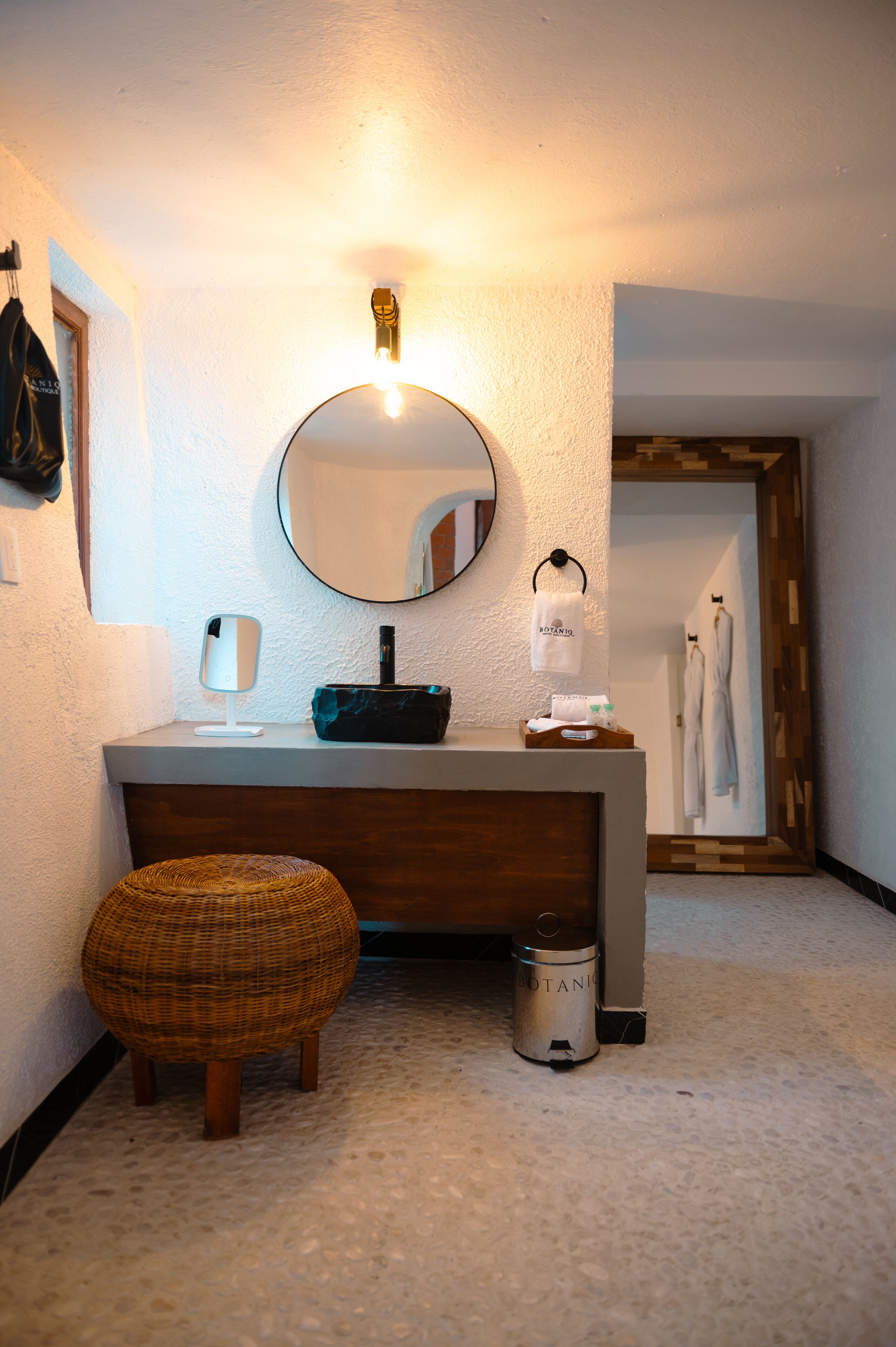 Ein Badezimmer mit rundem Spiegel, Waschbecken, Spender für Handtücher und einem kleinen Hocker.