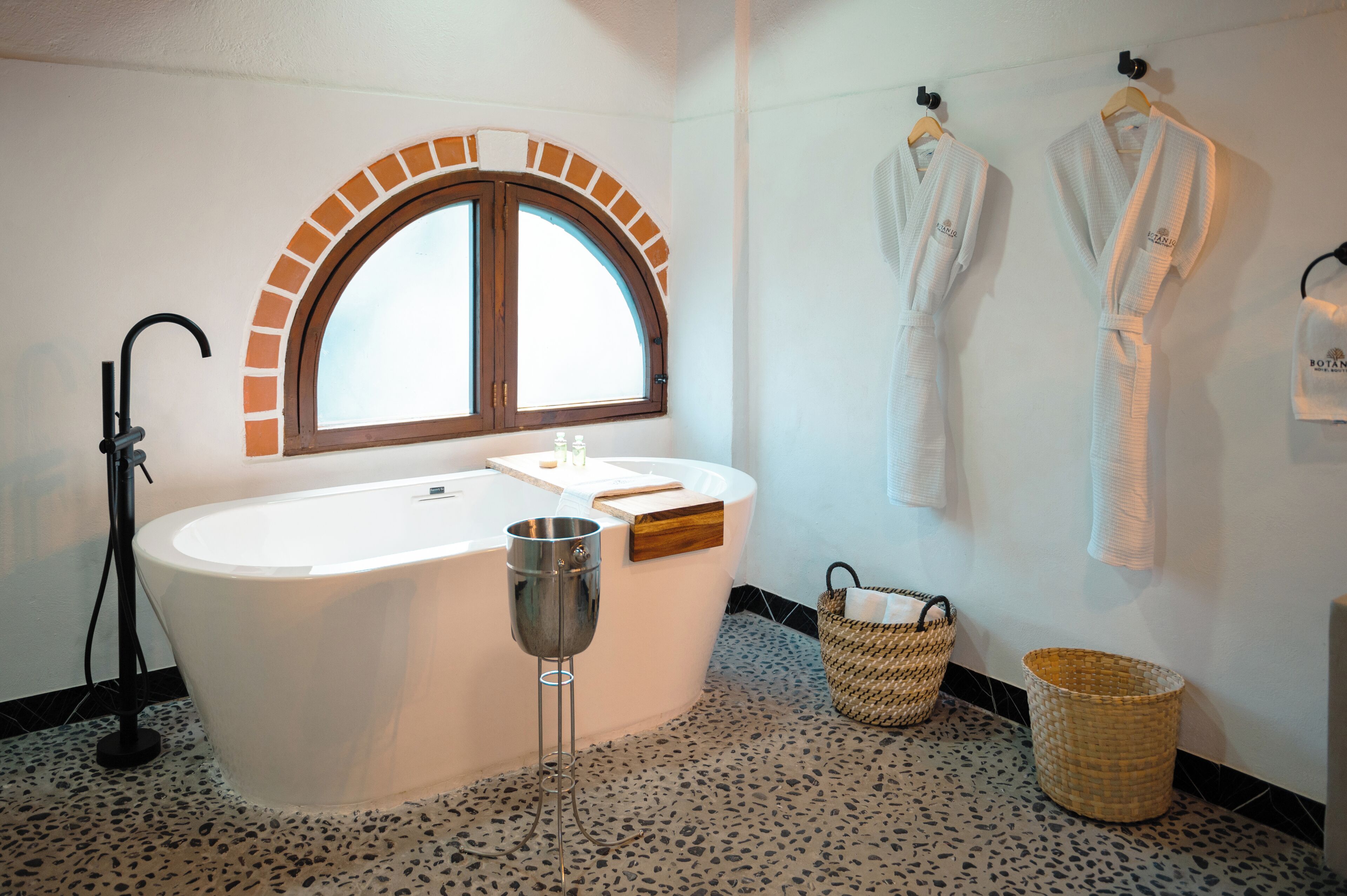 Un baño moderno con una bañera independiente, un ventanal con diseño de ladrillo en arco y dos batas blancas colgadas en ganchos.