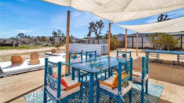 An outdoor dining area with a glass table and blue chairs, shaded by a large white canopy, set against a desert landscape with cacti and hills. - Stargazer Ranch - Stunning 5 BR + Hot Tub, Pool, Fire Pit (Yucca Valley)