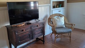 A wooden TV stand with a flat-screen TV, a wicker chair with a cushion, and a shelf with decorative items. - Cozy Home on Vine Street  (Paso Robles)