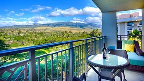 Ein Balkon mit Glastisch und Korbstühlen, mit Blick auf eine Berglandschaft mit Grünflächen und Gebäuden.