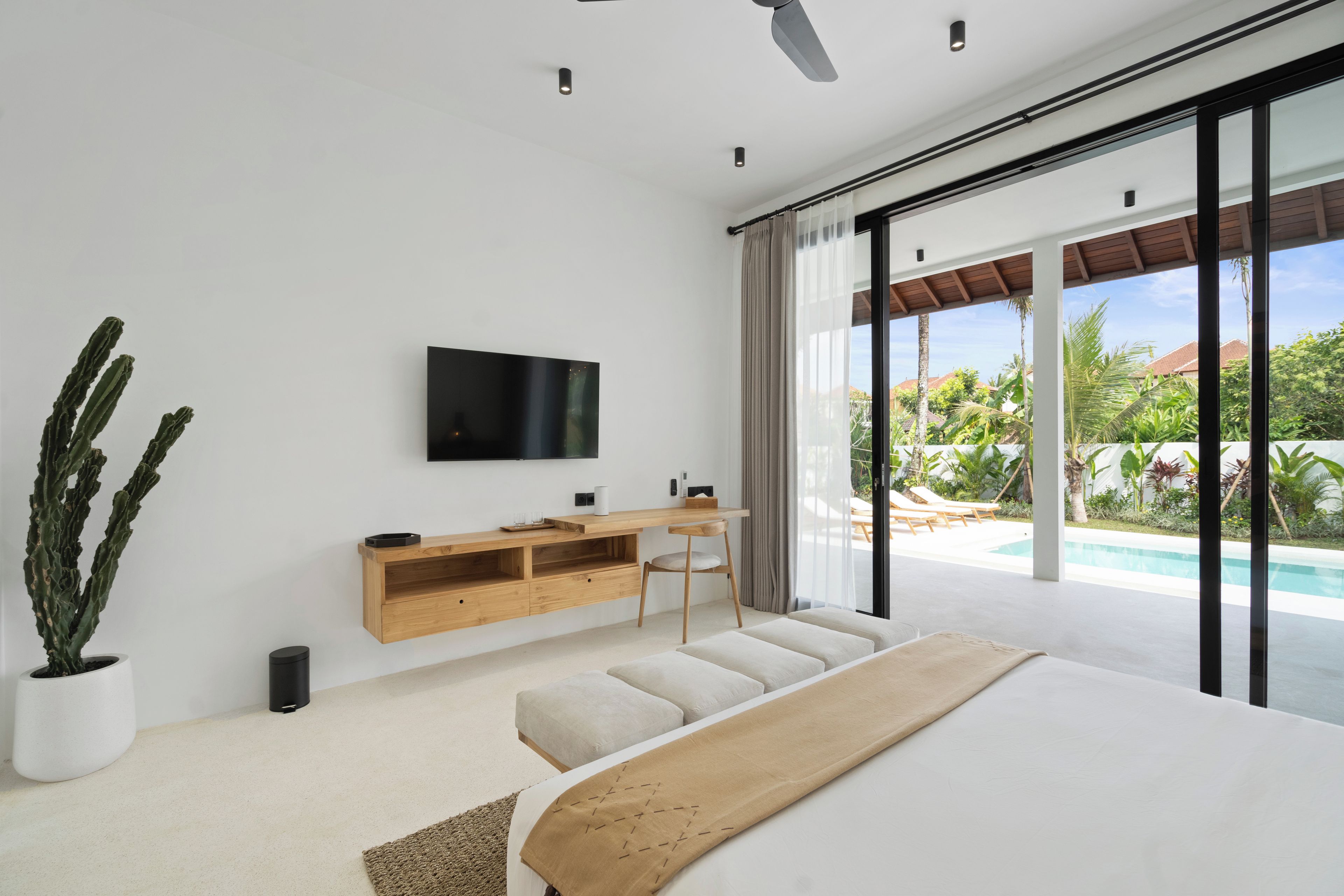 Quarto moderno com uma cama grande, TV na parede, mesa de console de madeira e vista para uma piscina e vegetação lá fora.