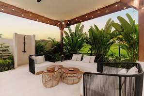 An outdoor seating area with a sofa, chairs, and round tables, surrounded by lush greenery and a view of a field. - Marbella Villa by DH (Ubud)