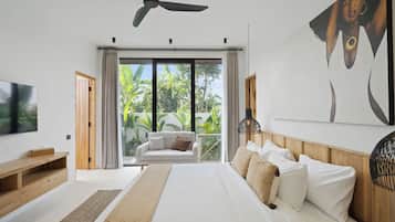A modern bedroom with a large bed, a ceiling fan, a TV, and a view of greenery through the window.