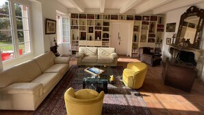Un salon spacieux avec un canapé beige, un fauteuil jaune et une table basse. On y trouve des étagères remplies de livres et d’objets décoratifs.