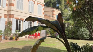 Un bâtiment doté d’un toit en tuiles et de volets blancs, entouré d’un jardin bien entretenu où poussent différentes espèces de plantes et où s’étend une pelouse.