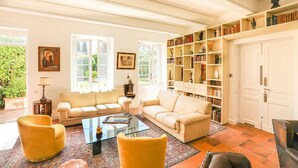 Un salon spacieux avec un canapé beige, des fauteuils jaunes, une table basse en verre et une grande bibliothèque remplie de livres et d’objets décoratifs.