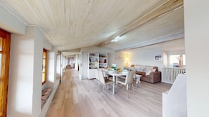 En rummelig, åben stue med højt til loftet og synlige træbjælker. Der er et spiseområde med et rundt bord og stole, et siddeområde med en sofa, og en indbygget reol.