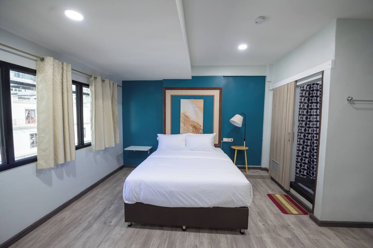 A hotel room with a large bed, a bedside table, a lamp, and a view of the outside through a window with curtains.