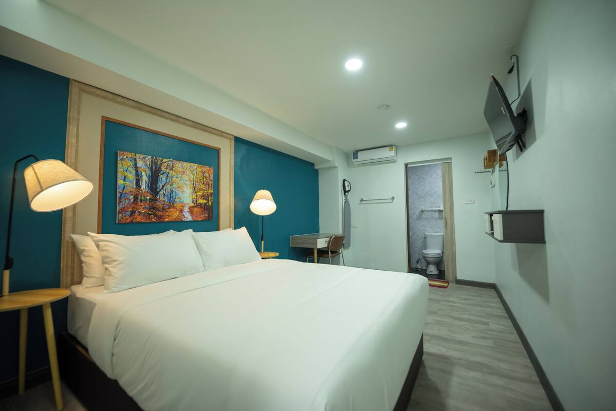 A hotel room with a bed, two bedside lamps, a painting, a desk, a chair, a TV, an air conditioning unit, and a bathroom with a toilet and shower.