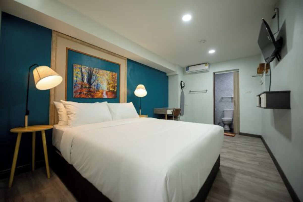 A hotel room with a large bed, two bedside lamps, a painting on the wall, a television mounted on the wall, and a door leading to a bathroom.