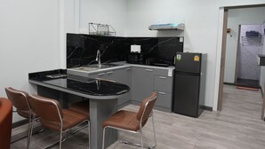 Une cuisine compacte dotée d’un plan de travail en marbre noir, d’une cuisinière encastrée et d’un réfrigérateur. Deux chaises marron sont disposées autour d’une petite table à manger.
