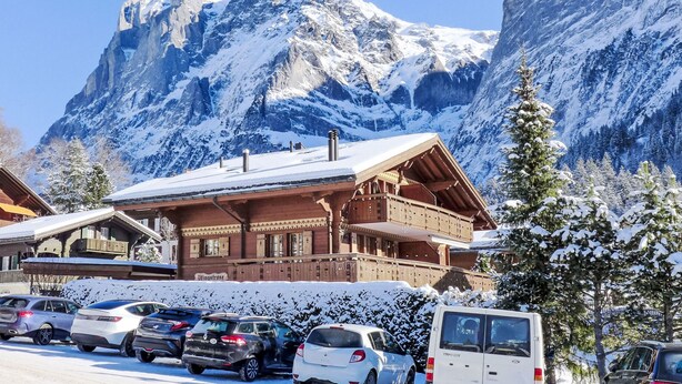 A mountain lodge with a snow-covered roof, parked cars, and a view of a snow-capped mountain.