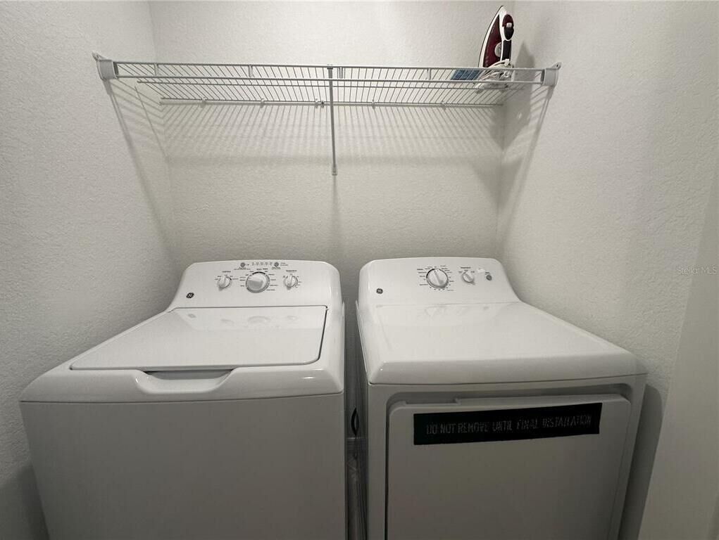Two white front-loading washing machines in a laundry area with a metal shelf above them.