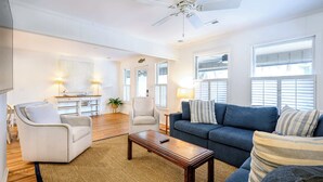 A spacious living room with a white armchair, a blue sofa, and a wooden coffee table. There is a ceiling fan, large windows with blinds, and a small table with a lamp. - 1 Block to Beach - Huge Screen Porch & Lagoon view (Edisto Island)