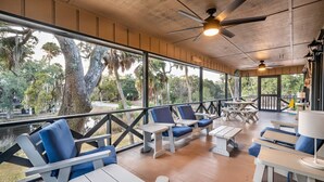 A covered outdoor seating area with wooden tables and blue cushioned chairs, overlooking a natural landscape with trees and a body of water. - 1 Block to Beach - Huge Screen Porch & Lagoon view (Edisto Island)