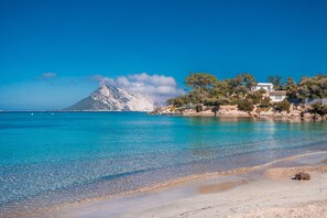 A coastal landscape with a clear blue sea, a sandy beach, and a mountain in the background.