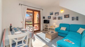 A living room with a blue sofa, a television on a white stand, a dining table with chairs, and a large window with curtains. - Quiet Sanibel, Monte Petrosu, Italy (Monte Petrosu)