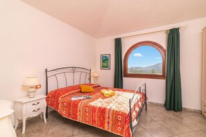Ein Schlafzimmer mit Bett, Nachttisch, einem Fenster mit Vorhängen und Blick ins Freie.